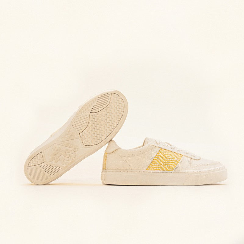 Paire de baskets basses en toile beige avec un motif ethnique jaune brodé sur les côtés.
Une chaussure est posée à plat tandis que l'autre est inclinée pour dévoiler la semelle texturée avec le logo N'Go Shoes.