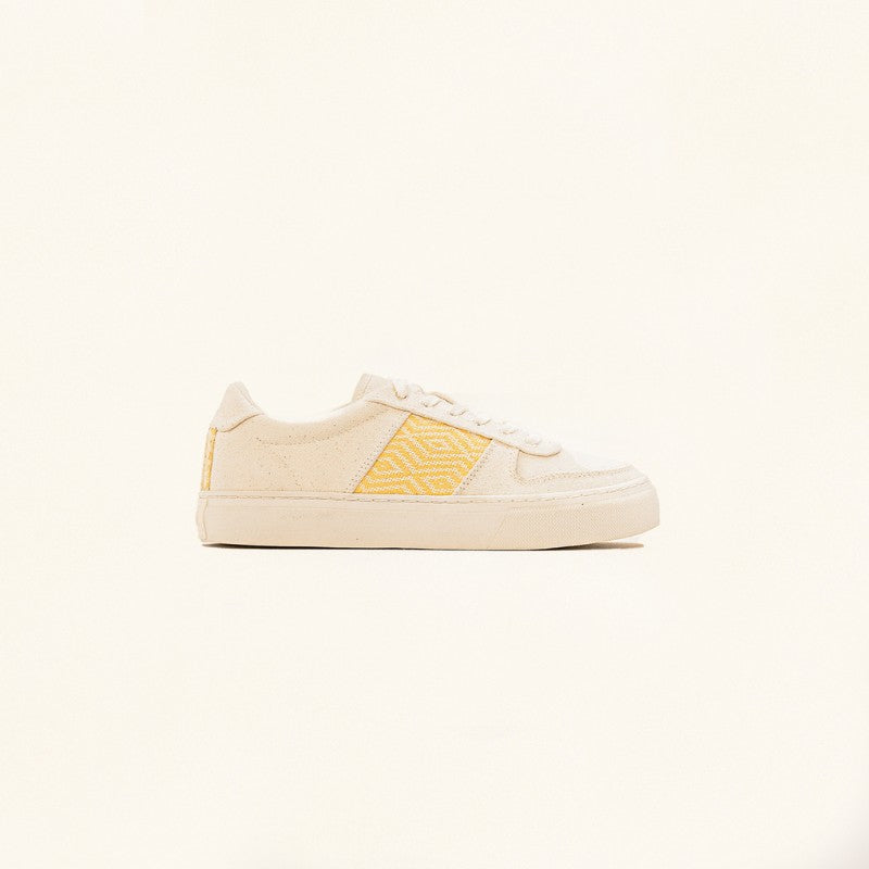 Vue latérale de la basket posée sur un fond clair, présentant son design épuré et ses détails artisanaux.
La silhouette minimaliste met en valeur la combinaison du tissu beige et des broderies jaunes