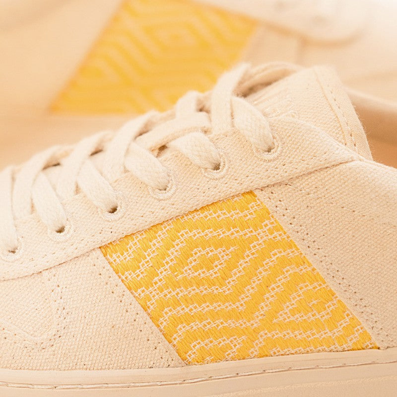 Zoom sur le côté de la basket, montrant de près le motif ethnique jaune brodé sur la toile beige.
Les lacets en coton beige et la texture du tissu naturel sont clairement visibles.