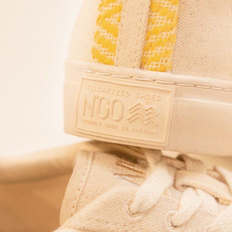 Gros plan sur l’arrière de la chaussure, mettant en valeur l’inscription en relief "N'GO Vulcanized Shoes, Proudly Made in Vietnam".
Le détail du tissu beige et du motif jaune brodé est visible le long du talon.